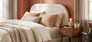 how to pick a bed buying guide banner - cosy bedroom with cream boucle fabric bed with soft orange and cream bedding, wooden bedside table, terracotta wall