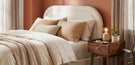how to pick a bed buying guide banner - cosy bedroom with cream boucle fabric bed with soft orange and cream bedding, wooden bedside table, terracotta wall