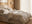 modern softly lit bedroomw with cream fabric double bed and comfy rumpled layered bedding, round wooden bedside table, accent chair with jacket draped over it