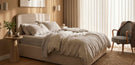 modern softly lit bedroomw with cream fabric double bed and comfy rumpled layered bedding, round wooden bedside table, accent chair with jacket draped over it