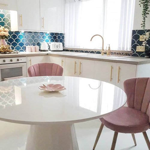 birght kitchen-diner. Round white gloss table with tapered central plinth, 2 pink velvet chairs. Petrol blue tiling to walls, white cabinet.
