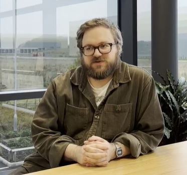 Arthur, Marketplace Executive, white male with fair hair and beard with glasses, in khaki shirt, sat at table talking. 