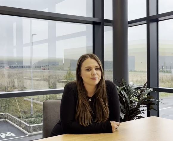 Hattie Marketing Manager headshot, white woman long brown hair, in back top, sat at table smiling