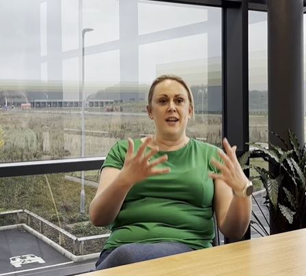 Mel HR Manager, white woman, blonde hair tied back, in green t-shirt, sat at table talking and smiling