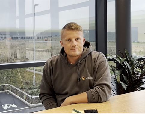 Zak Systems Administrator, white male, close cropped blonde hair, in grey Furniturebox hoodie, sat at table, smiling