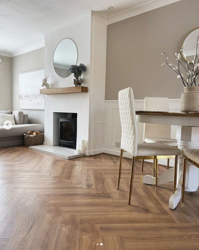 living dining room wiith warm neutrals to illustrate paint makeover. round wooden dining table with white and gold chairs, wooden mantle over fireplace and grey sofa