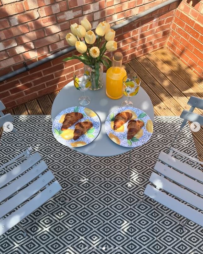 grey 2 seat metal outdoor table and chairs in corner of brick outdoor space, wth orange juice and croissants on table